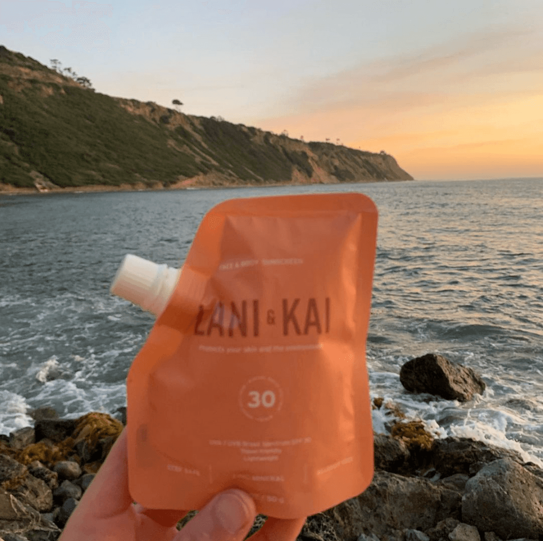 A Lani & Kai sunscreen pouch held up against the ocean and coastline at sunset